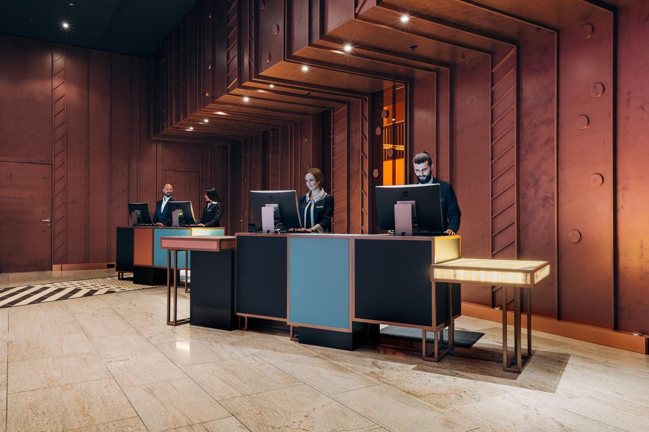 Hotel Pullman Berlin Schweizerhof Exterior foto The image shows a modern hotel lobby reception area. There are several reception desks made of dark materials with illuminated accents. Behind the desks, three people are standing, likely staff members, engaged with computer screens. The lobby featur
